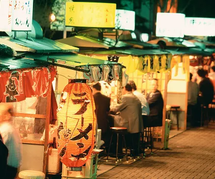 夜の賑わう屋台街の風景。手前に赤い提灯が灯り、「ラーメン」と書かれている。奥には複数の屋台が連なり、黄色い看板が光っている。