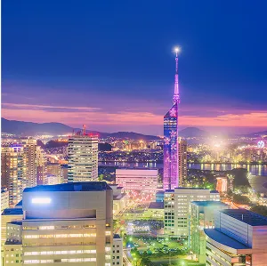 夕暮れ時の都市の風景。中央にピンク色にライトアップされた高層タワー（福岡タワー）がそびえ立ち、その周囲には複数の高層ビルが建ち並んでいる。