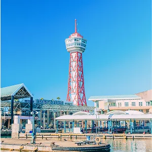 青空の下、赤と白の鉄骨で組まれた特徴的な形のタワーがそびえ立つ。タワーの周辺には、白い建物と緑色の屋根の建物が並んでいる。手前には水面が広がり、小型のボートが浮かんでいる。
